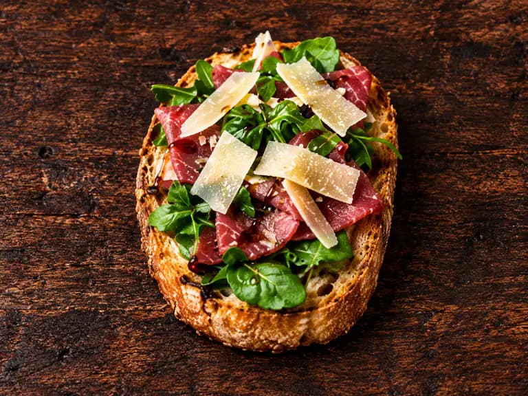 Bruschette con carpaccio di manzo con la farcitura su pane casereccio tostato, fotografate su un piano di legno scuro