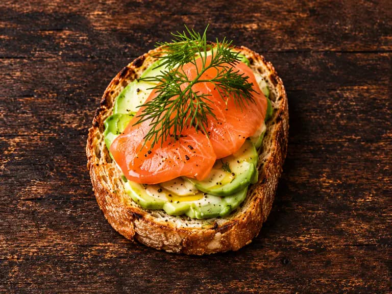Bruschette con salmone e avocado con la farcitura su pane casereccio tostato, fotografate su un piano di legno scuro