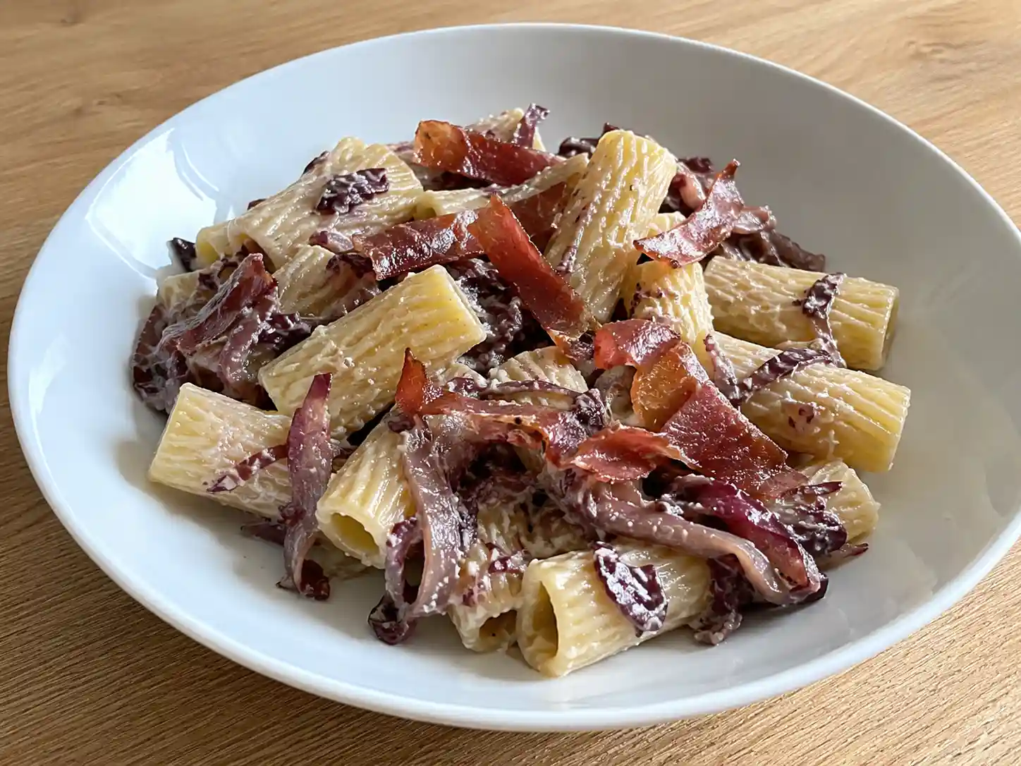 Pasta al radicchio e speck in ciotola bianca con rigatoni cremosi, radicchio stufato e speck dorato