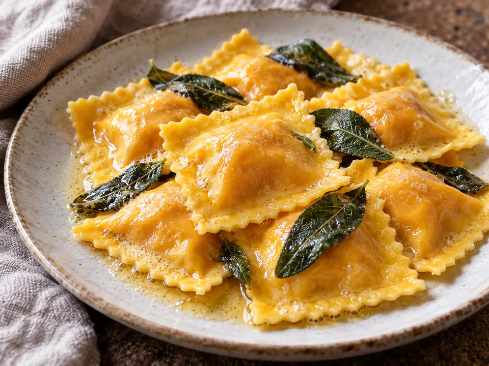 Ravioli di zucca quadrati con burro fuso e foglie di salvia su un piatto chiaro in luce naturale