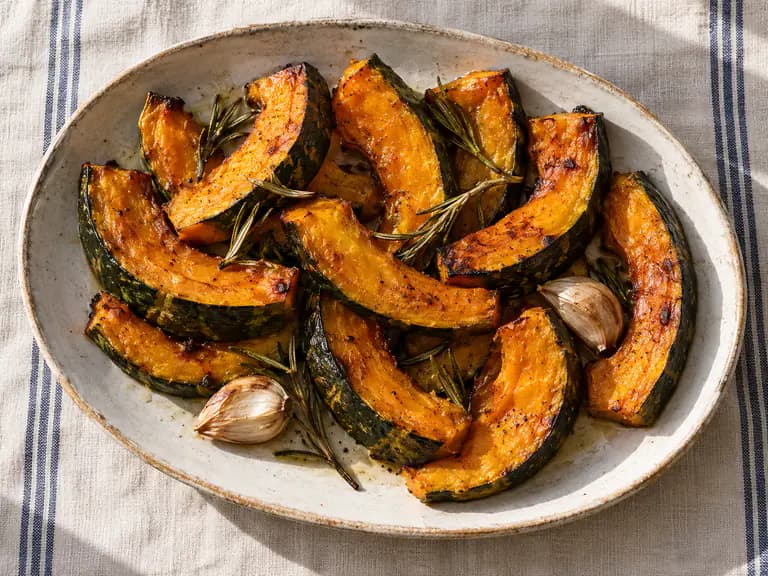 Spicchi di zucca Delica arancione con bordi dorati su una teglia, rosmarino e aglio in camicia